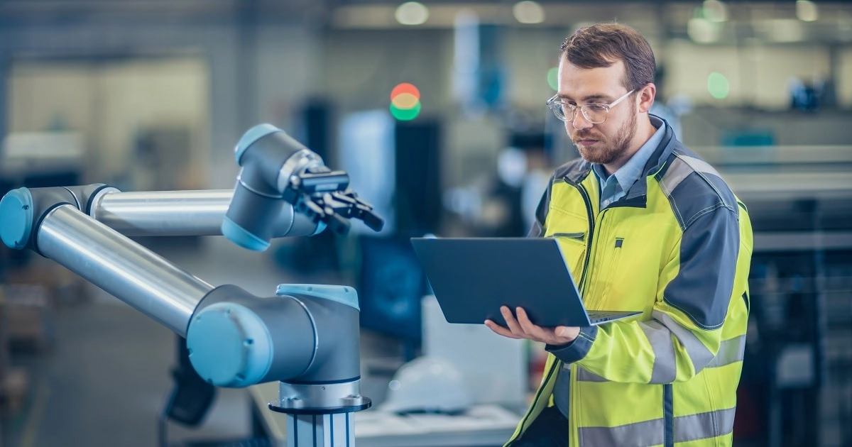 6654c5b9-0e4d-4540-bdf6-c0f9aaeb81db_Industrial_manufacturing_article-min A man in a yellow vest is seated, working intently on a laptop
