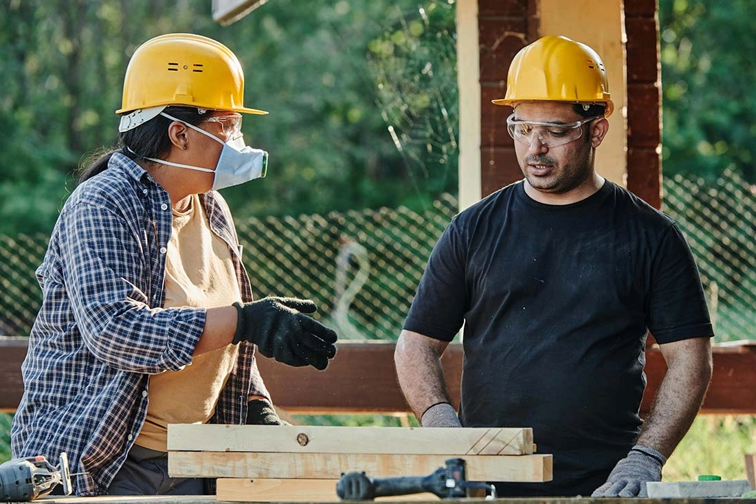 pexels-mikael-blomkvist-8961619-compressed Two people wearing hard hats and safety glasses work together at a wooden table, engaged in a construction task