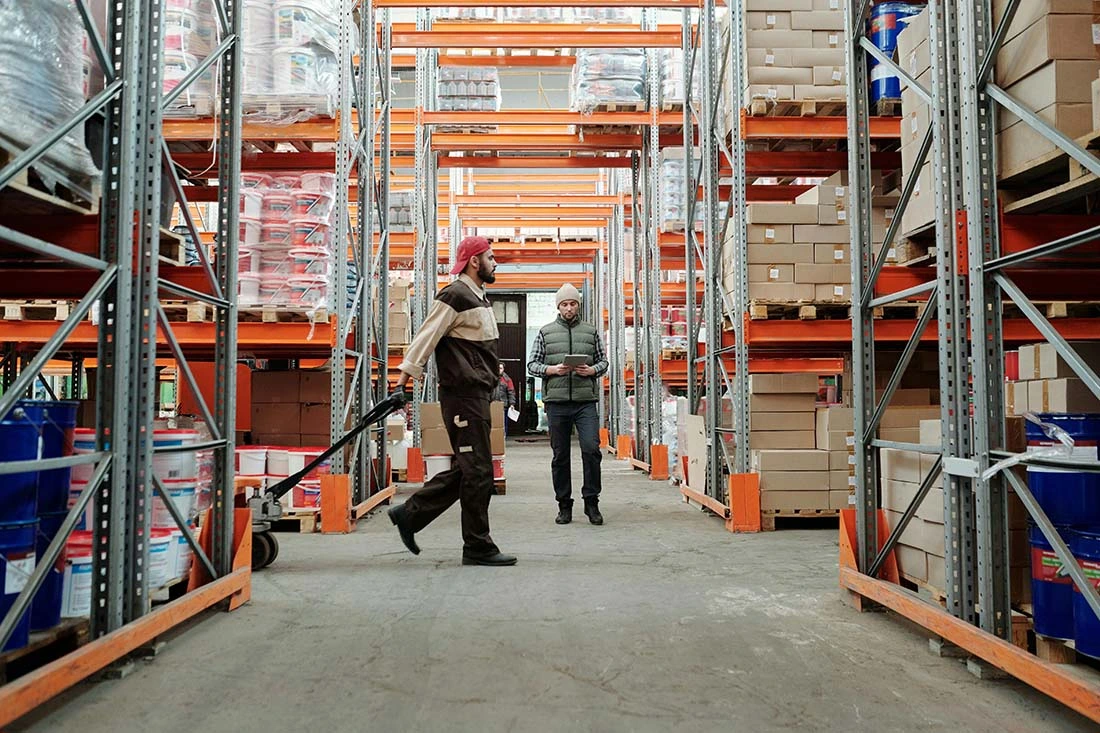 pexels-tiger-lily-4487365-compressed Two individuals stroll through a spacious warehouse, flanked by shelves filled with products